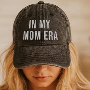 Könnte beinhalten: Eine anthrazitfarbene Baseballkappe mit verwaschenem, abgenutztem Finish. Auf der Vorderseite befindet sich der gestickte weiße Schriftzug "IN MY MOM ERA" in einer klaren serifenlosen Schrift. Die Kappe hat eine strukturierte Krone und einen Schirm.