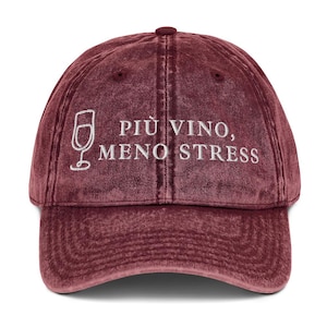 Op de afbeelding: Bordeauxkleurige baseballcap met de geborduurde tekst "PIÙ VINO, MENO STRESS" in het wit. De cap heeft een wijnglasafbeelding en een gebogen klep. De hoed is gemaakt van een zachte, getextureerde stof.