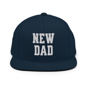 Könnte beinhalten: Eine marineblaue Snapback-Cap mit dem in weißen Blockbuchstaben auf die Vorderseite gestickten Text NEW DAD. Die Mütze hat eine strukturierte Krone, einen flachen Schirm und einen Knopf an der Oberseite, was ein klares und schlichtes Design ergibt.