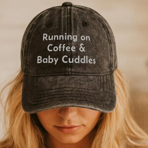 May include: A charcoal grey baseball cap with a distressed, washed-out finish. The front of the cap features white embroidered text that reads Running on Coffee & Baby Cuddles. The hat has a structured crown, a curved brim, and a top button.