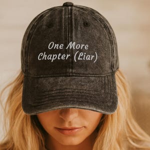 Könnte beinhalten: Eine anthrazitfarbene Baseballkappe mit verwaschener Optik. Auf der Vorderseite befindet sich eine weiße Stickerei mit dem Text One More Chapter (Liar) in einer klaren, serifenlosen Schrift. Die Kappe verfügt über eine strukturierte Krone und einen Schirm.