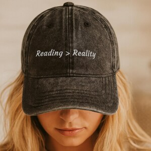 Könnte beinhalten: Eine anthrazitfarbene Baseballkappe mit verwaschener Optik. Auf der Vorderseite befindet sich der in Weiß gestickte Schriftzug Reading > Reality in einer serifenlosen Schrift, der mittig über dem gebogenen Schirm platziert ist.