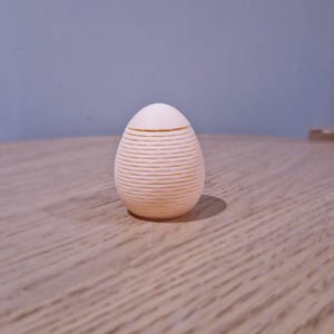Può includere: Un oggetto decorativo a forma di uovo, di colore pesca chiaro, con dettagli orizzontali a coste. La parte superiore è un cappuccio liscio e arrotondato. L'oggetto è posto su una superficie di legno.