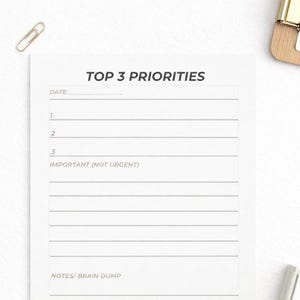 May include: A white Top 3 Priorities planner with lined sections for date, priorities, and notes. The planner includes the text "TOP 3 PRIORITIES" and "TOP 3 PRIORITY PLANNER". A silver pen and paperclip are also visible.