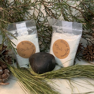 May include: Two clear plastic bags filled with white granules, labeled "Blessed Eggshells by Nature 1 oz." A dark gray stone sits between the bags, with pine needles and evergreen branches in the background.