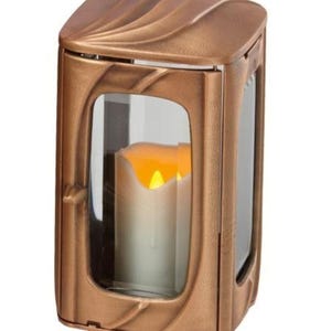 Pode incluir: Uma lanterna de cor bronze com formato retangular e parte superior curva. A lanterna possui painéis de vidro transparente e uma vela falsa no interior, emitindo um brilho quente e amarelo. O design apresenta linhas decorativas e fluidas.