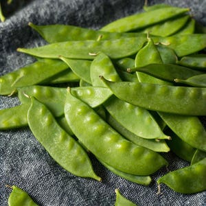 May include: A pile of fresh, vibrant green snap peas. The pods are smooth, with a slightly bumpy texture, and are arranged on a dark gray, textured surface. The peas are a bright, natural green color, suggesting freshness.