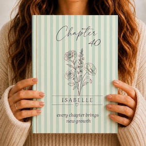 Peut inclure: Un livre à couverture rigide avec des rayures verticales bleu clair et blanches. La couverture présente le texte "Chapter 40", un dessin au trait d'un bouquet floral, le nom "ISABELLE" avec un petit cœur et "every chapter brings new growth".