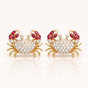 May include: A pair of gold-colored crab earrings. The crab's body is encrusted with small, clear gemstones, and the claws are adorned with red gemstones. The earrings have a polished finish and are set against a white background.