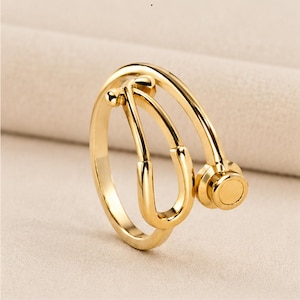 May include: A gold-colored ring in the shape of a stethoscope. The ring features the curved tubes and the circular chest piece of a stethoscope. The ring is made of a shiny, reflective metal and is set against a neutral background.