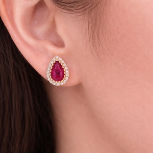 Op de afbeelding: Een gouden oorbel in de vorm van een druppel met een levendige rode middensteen, omringd door een halo van kleine, sprankelende diamanten. De oorbel wordt getoond tegen de achtergrond van het oor en de huid van een persoon, wat het elegante ontwerp van de sieraden benadrukt.