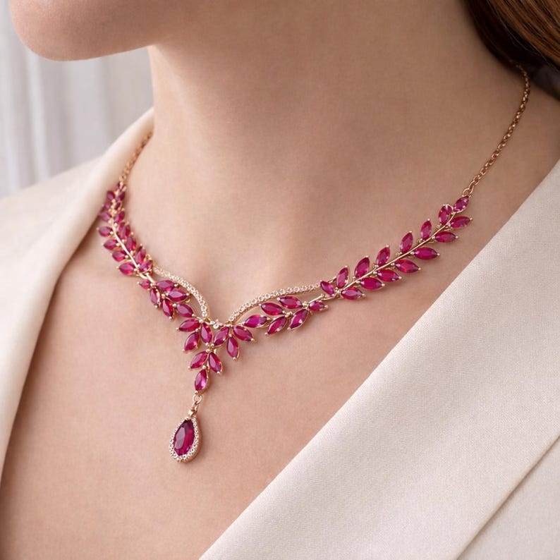 May include: A rose gold necklace featuring a leaf design with marquise-cut ruby-colored gemstones. The necklace has a pendant with a teardrop-shaped ruby surrounded by small white gemstones. A delicate and elegant piece of jewelry.
