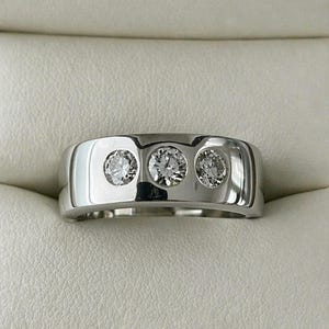 May include: A polished, silver-toned ring featuring three round, clear gemstones set in a horizontal row. The ring has a wide band with a smooth, reflective surface. The gemstones sparkle against the light background.
