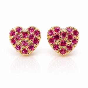 Peut inclure: Une paire de boucles d'oreilles en forme de cœur. Chaque boucle d'oreille est dorée et incrustée de nombreuses petites pierres précieuses roses rondes. Les boucles d'oreilles sont présentées sur fond blanc.