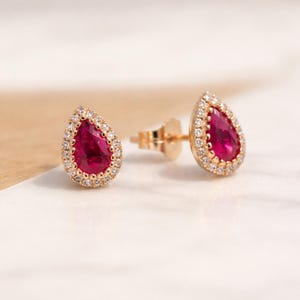 May include: A pair of gold stud earrings featuring pear-shaped, vibrant red gemstones surrounded by a halo of small, sparkling white stones. The earrings are set against a soft, neutral background, highlighting their elegant design.