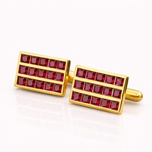 May include: A pair of gold cufflinks with a rectangular design. Each cufflink features a grid of square, deep red gemstones. The cufflinks are set against a white background, highlighting their polished finish.