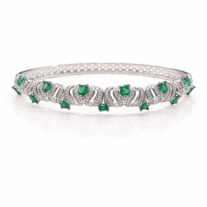 May include: A silver bracelet with a repeating pattern of emerald-green gemstones and small clear stones. The bracelet has a curved design with the gemstones set in a decorative, wave-like pattern. The bracelet is displayed on a white background.