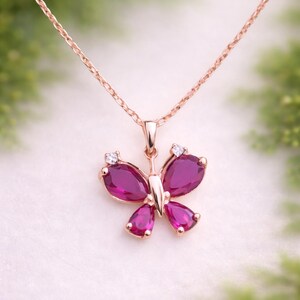 May include: Rose gold butterfly pendant necklace with a delicate chain. The butterfly pendant features vibrant, teardrop-shaped, magenta-coloured gemstones and small, clear accent stones. The necklace is displayed against a soft, blurred background.