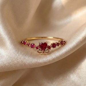 Peut inclure: Une bague dorée avec une pierre centrale en forme de cœur rouge et des accents de rubis plus petits. La bague présente un motif de nœud sous le cœur, sur un tissu crème.