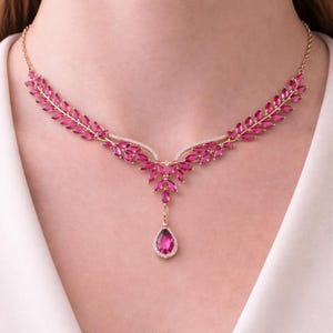 May include: A gold-toned necklace featuring a leaf-like design with vibrant pink gemstones. The necklace has a central pendant with a teardrop-shaped pink stone, and is set against a white background.