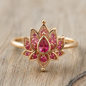 Peut inclure: Une bague en or arborant un motif central en forme de fleur de lotus. Les pétales sont ornés de multiples pierres précieuses roses rondes et d'une pierre rose en forme de poire au centre. L'anneau en métal est lisse et poli, avec un motif floral.