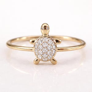 Op de afbeelding: Een goudkleurige ring met een schildpadontwerp. De schildpadschild is bezet met kleine, sprankelende steentjes. De ring heeft een dunne, gepolijste gouden band. De schildpad heeft kleine, afgeronde pootjes en een kop met twee kleine, heldere steentjes als ogen.