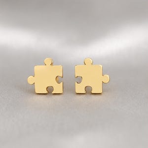 May include: A pair of gold-toned stud earrings shaped like puzzle pieces. Each earring features a smooth, polished metallic surface with the characteristic interlocking tabs and blanks of a jigsaw puzzle piece, designed for earlobe placement.