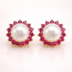May include: A pair of stud earrings featuring a large, round, white pearl center surrounded by a halo of small, vibrant red gemstones. The earrings have a gold-colored setting and a polished finish.