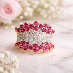 May include: A wide gold ring with a central band of small, clear stones, framed by rows of vibrant red gemstones. The ring's design is set against a soft, blurred background with pink and white flowers, creating a luxurious aesthetic.