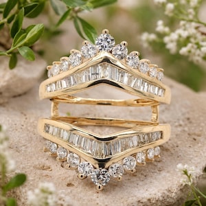 May include: A gold-colored ring guard with a V-shaped design. The ring is encrusted with multiple clear, sparkling diamonds, including round and baguette cuts. The ring guard is designed to enhance the look of an engagement ring.