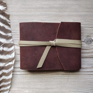 May include: A dark brown leather journal with a cream-colored tie closure. The journal is rectangular and appears to be handmade. A large feather with brown and white stripes is next to the journal on a wooden surface.