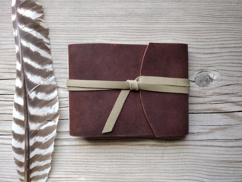 May include: A dark brown leather journal with a cream-colored tie closure. The journal is rectangular and appears to be handmade. A large feather with brown and white stripes is next to the journal on a wooden surface.