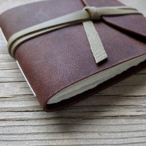 May include: A brown leather journal with a cream-colored strap tied around it. The journal's pages are white, and the cover has a textured appearance. The journal is closed and resting on a wooden surface.