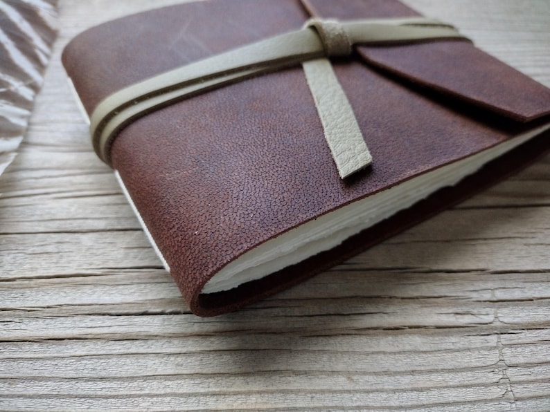 May include: A brown leather journal with a cream-colored strap tied around it. The journal's pages are white, and the cover has a textured appearance. The journal is closed and resting on a wooden surface.