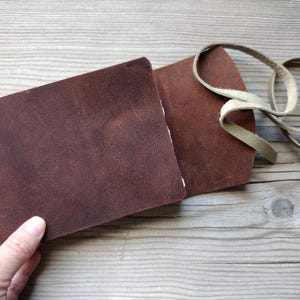 May include: A brown leather wallet with a cream-colored leather strap. The wallet is open, revealing the interior. The leather has a textured appearance. The wallet is on a wooden surface.