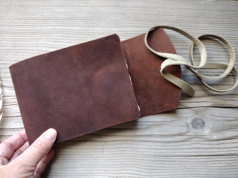 May include: A brown leather wallet with a cream-colored leather strap. The wallet is open, revealing the interior. The leather has a textured appearance. The wallet is on a wooden surface.