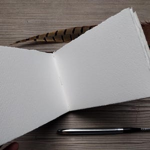 May include: An open, blank watercolor sketchbook with textured white pages. A silver and black paintbrush lies next to the open book. A brown and black feather and a leather strap are also visible.