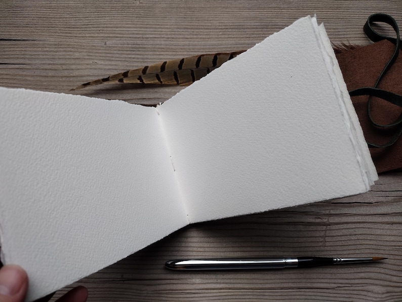 May include: An open, blank watercolor sketchbook with textured white pages. A silver and black paintbrush lies next to the open book. A brown and black feather and a leather strap are also visible.