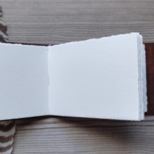 May include: Open journal with blank white pages and a brown leather cover. The journal is held open, revealing the textured paper. A brown leather strap and a decorative feather are also visible.