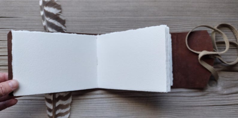 May include: Open journal with blank white pages and a brown leather cover. The journal is held open, revealing the textured paper. A brown leather strap and a decorative feather are also visible.