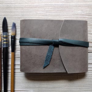 May include: A gray leather journal with a dark green leather tie, next to three paint brushes. The brushes have wooden handles and black bristles. The journal is rectangular and has a soft, suede-like texture.
