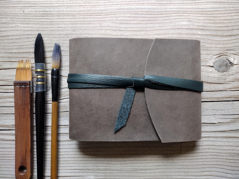 May include: A gray leather journal with a dark green leather tie, next to three paint brushes. The brushes have wooden handles and black bristles. The journal is rectangular and has a soft, suede-like texture.