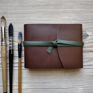 May include: A brown leather journal with a green tie closure, next to three paint brushes with wooden handles and black bristles. The brushes vary in size and bristle shape, suggesting different painting techniques. The journal is closed and appears to be new.