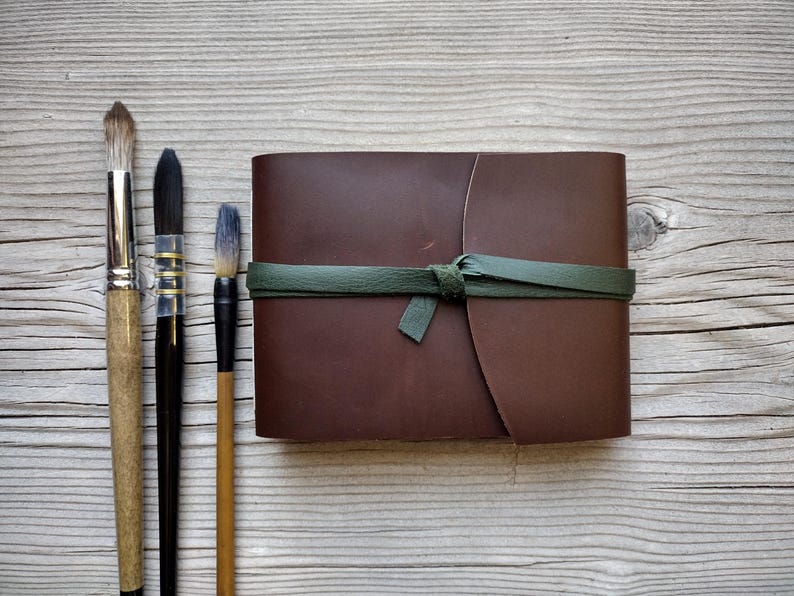 May include: A brown leather journal with a green tie closure, next to three paint brushes with wooden handles and black bristles. The brushes vary in size and bristle shape, suggesting different painting techniques. The journal is closed and appears to be new.