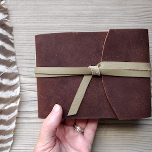May include: A dark brown leather journal with a light green leather tie closure. The journal is rectangular and has rounded edges. A large feather with brown and white stripes is next to the journal.