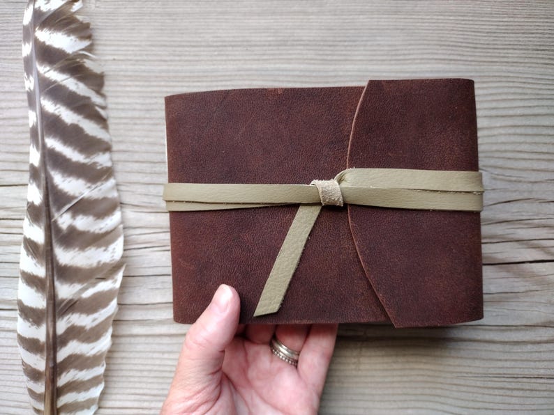 May include: A dark brown leather journal with a light green leather tie closure. The journal is rectangular and has rounded edges. A large feather with brown and white stripes is next to the journal.