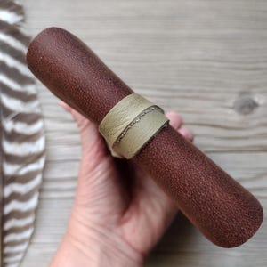 May include: A cylindrical brown leather item with a textured surface, secured by a light green leather band. A feather is visible in the background. The item appears to be a pen or pencil case.