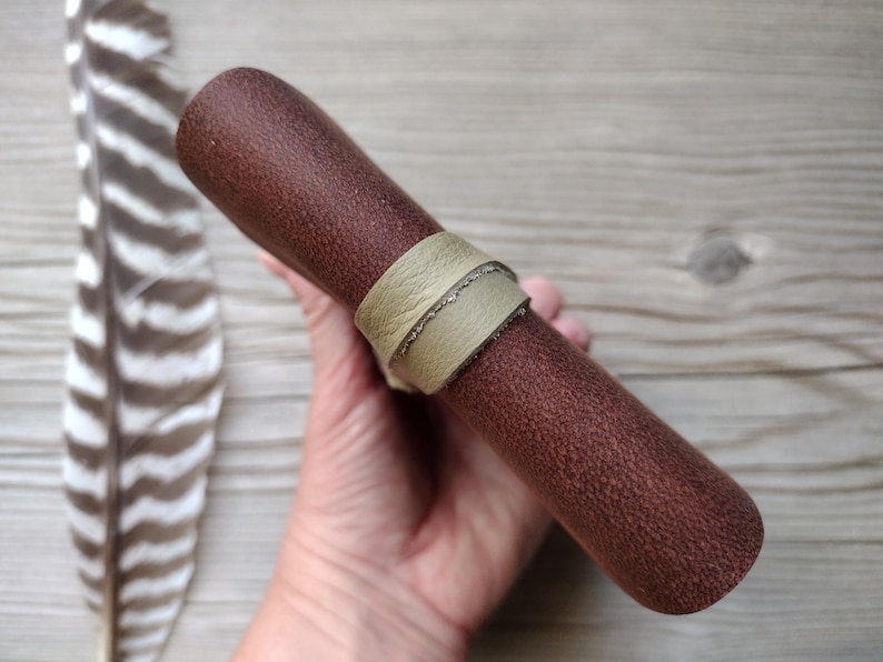May include: A cylindrical brown leather item with a textured surface, secured by a light green leather band. A feather is visible in the background. The item appears to be a pen or pencil case.