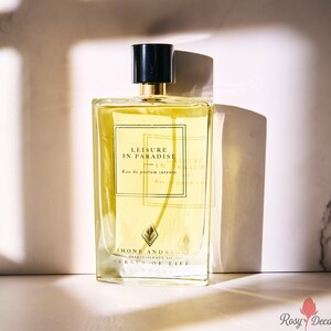 K&ouml;nnte beinhalten: Ein rechteckiger Parf&uuml;mflakon mit schwarzem Verschluss und goldenen Akzenten. Die Flasche enth&auml;lt eine gelbe Fl&uuml;ssigkeit und tr&auml;gt den Text "LEISURE IN PARADISE" und "SIMONE ANDREOLI". Der Hintergrund ist eine Marmoroberfl&auml;che mit Schatten.