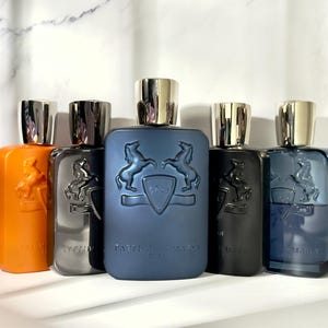 May include: A collection of perfume bottles in various colors, including orange, gray, black, and blue. Each bottle has a silver cap and a horse emblem. The bottles are arranged in a row, showcasing a range of fragrances.